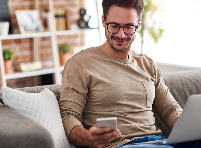 Hombre en un sofá mirando su teléfono mientras tiene un portátil en su regazo